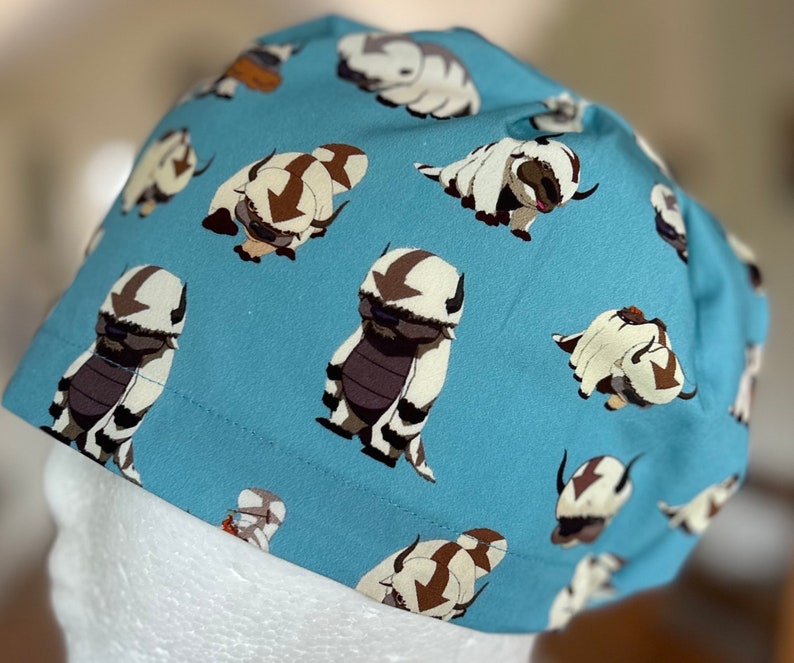 Peut inclure: Un chapeau en tissu bleu avec un motif r&eacute;p&eacute;t&eacute; d'animaux de dessin anim&eacute; blancs et bruns. Les animaux sont stylis&eacute;s et ressemblent &agrave; une cr&eacute;ature mythique avec une longue crini&egrave;re flottante.