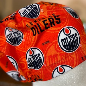 May include: Orange and black fabric scrub hat with a repeating pattern of the Edmonton Oilers logo. The logo features a stylized oil drop with the word "Oilers" inside a circle. The text "Edmonton Oilers" is also visible on the hat.