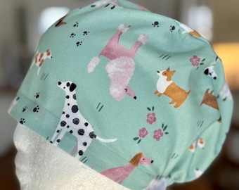 Könnte beinhalten: Eine helltürkisfarbene OP-Haube mit einem sich wiederholenden Muster verschiedener Hunderassen, darunter ein Dalmatiner, ein Pudel und ein Corgi. Das Design umfasst auch Pfotenabdrücke und kleine rosa Blumen.