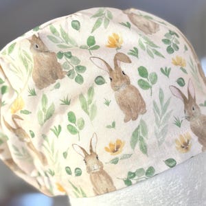 May include: A white scrub cap featuring a repeating pattern of brown rabbits, green leaves, and yellow flowers. The cap is made of a soft fabric and is designed to cover the head.