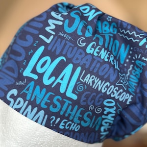 May include: A blue and black scrub hat with a variety of medical terms printed on it in white. The terms include "induction", "intubation", "local", "general", "anesthesia", "spinal", "laryngoscope", "echo", "nausea", and "cool me".