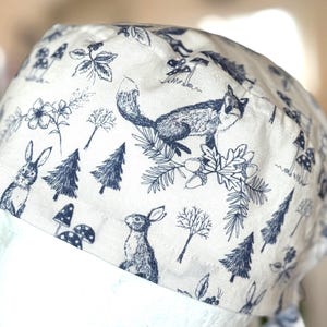 Puede incluir: Un gorro quirúrgico blanco y azul marino estampado con un diseño de bosque. El gorro muestra ilustraciones de un zorro, conejos, árboles, setas y elementos florales. Hecho de tela suave y transpirable.