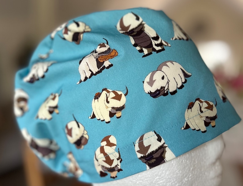Peut inclure: Un bonnet de chirurgien bleu avec un motif r&eacute;p&eacute;t&eacute; de bisons de dessin anim&eacute; blancs et bruns. Les bisons sont dans diff&eacute;rentes poses, y compris debout, marchant et couch&eacute;s.