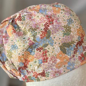 May include: A pink and white floral print scrub hat with a gathered top. The hat has a variety of flowers in shades of pink, blue, and orange.