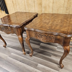 May include: Two antique wooden side tables with ornate carved details and curved legs. The tables have a dark brown finish and a patterned tabletop. The tables are placed on a gray and white wooden floor.