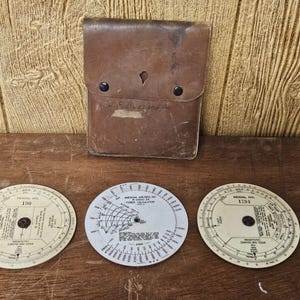 May include: Vintage leather case with three circular slide rules. The case is brown with a flap closure and two black snaps. The slide rules are off-white with black markings and dials. The image is on a wooden surface.