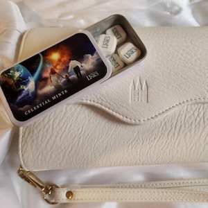 May include: A white leather clutch purse with a gold chain strap and a small tin of Celestial Mints with a colorful image of a family standing on a cloud with the Earth in the background. The tin has the words "Celestial Mints" and "Links of Love" printed on it.