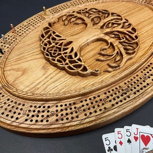 May include: An oval wooden cribbage board featuring a carved tree of life design in the center. The board includes rows of drilled holes for scoring and a set of metal pegs in various metallic finishes. A small hand of playing cards is visible.
