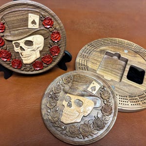May include: Two circular wooden cribbage boards featuring a carved skull wearing a top hat with an ace of spades card tucked into the band, surrounded by a wreath of roses. One board has red painted roses, while the other has natural wood roses.