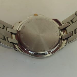 K&ouml;nnte beinhalten: Eine silberfarbene Armbanduhr mit Edelstahlarmband. Der Uhrenboden ist sichtbar und zeigt Text wie "WATER RESISTANT" und "ETA 955 114". Die Uhr hat eine goldfarbene Krone.
