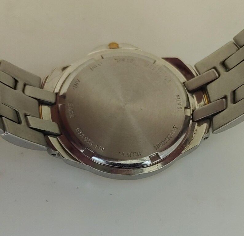 K&ouml;nnte beinhalten: Eine silberfarbene Armbanduhr mit Edelstahlarmband. Der Uhrenboden ist sichtbar und zeigt Text wie "WATER RESISTANT" und "ETA 955 114". Die Uhr hat eine goldfarbene Krone.