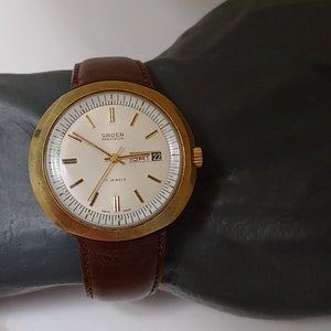 May include: A gold-toned Gruen Precision wristwatch with a white face, black hands, and a date window displaying "SAT 22". The watch has a brown leather band.