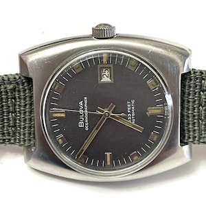 May include: A vintage Bulova Oceanographer watch with a dark gray face and a date display at the 3 o'clock position. The watch has a silver-tone case and a green fabric strap. The watch face has gold-tone hands and markers. The watch is marked "333 FEET AUTOMATIC".
