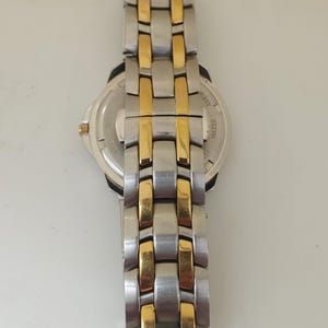 K&ouml;nnte beinhalten: Eine Edelstahl-Armbanduhr mit goldfarbenen Akzenten am Armband. Das Zifferblatt ist silberfarben mit schwarzen Details. Das Armband besteht aus rechteckigen Gliedern, die sich mit Silber und Gold abwechseln. Die Uhr ist wasserdicht.