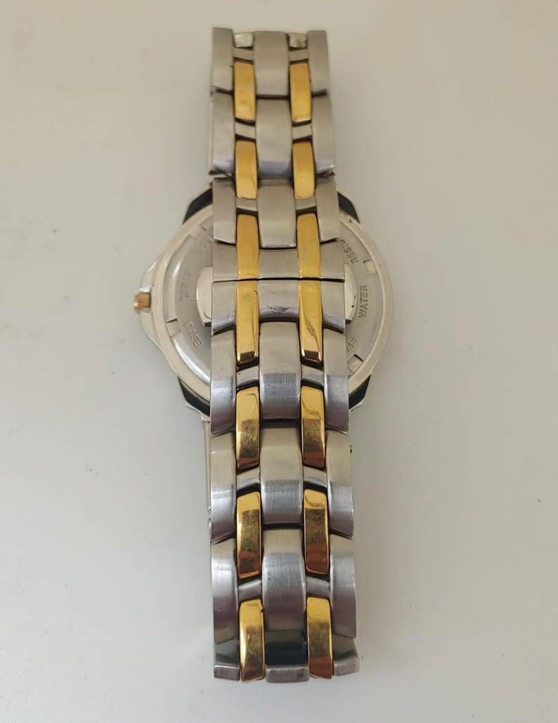 K&ouml;nnte beinhalten: Eine Edelstahl-Armbanduhr mit goldfarbenen Akzenten am Armband. Das Zifferblatt ist silberfarben mit schwarzen Details. Das Armband besteht aus rechteckigen Gliedern, die sich mit Silber und Gold abwechseln. Die Uhr ist wasserdicht.