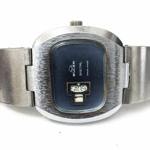 May include: A vintage Buler digital watch with a brushed silver-tone case and band. The watch face is dark blue with the brand name, "Buler Digital Swiss Made," and a date display showing "15" and "20".