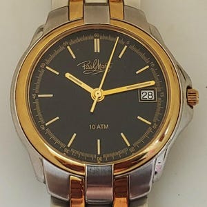K&ouml;nnte beinhalten: Armbanduhr mit schwarzem Zifferblatt, goldenen Zeigern und Datumsanzeige. Die Uhr hat eine goldene L&uuml;nette und Akzente mit einem silbernen Armband. Das Zifferblatt zeigt den Markennamen "Paul Marty" und den Text "10 ATM".