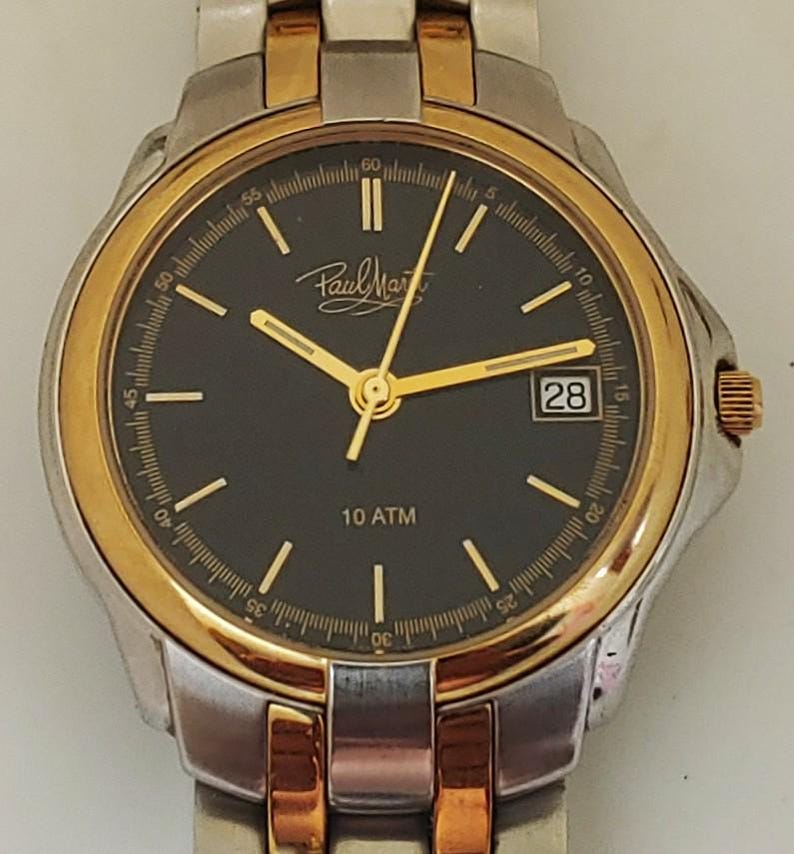 K&ouml;nnte beinhalten: Armbanduhr mit schwarzem Zifferblatt, goldenen Zeigern und Datumsanzeige. Die Uhr hat eine goldene L&uuml;nette und Akzente mit einem silbernen Armband. Das Zifferblatt zeigt den Markennamen "Paul Marty" und den Text "10 ATM".