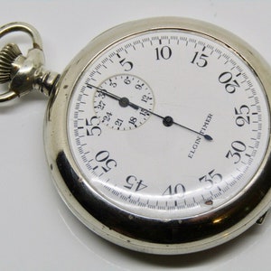 Pode incluir: Um relógio de bolso prateado com um mostrador branco e ponteiros pretos. O relógio tem um pequeno sub-mostrador e as palavras "ELGIN TIMER" impressas no mostrador.