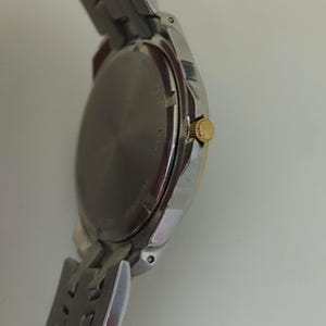 K&ouml;nnte beinhalten: Eine silberfarbene Armbanduhr mit goldfarbener Krone und silbernem Armband. Das Zifferblatt der Uhr ist dunkel, und das Armband besteht aus ineinandergreifenden Metallgliedern. Die Uhr ist abgewinkelt und zeigt die Seite und die R&uuml;ckseite.