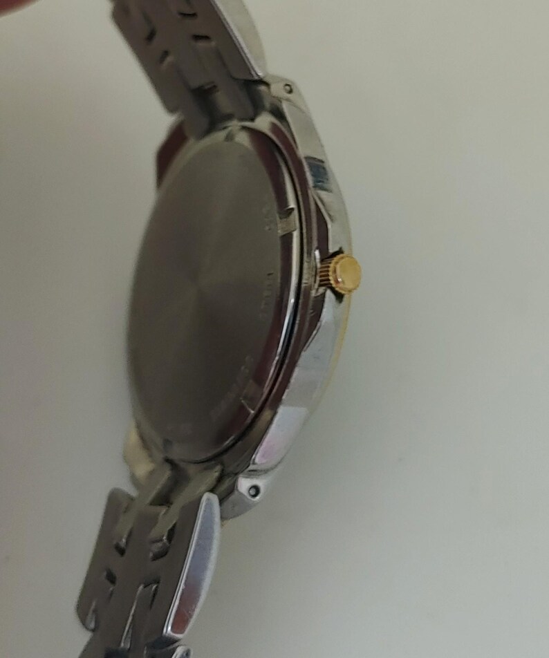 K&ouml;nnte beinhalten: Eine silberfarbene Armbanduhr mit goldfarbener Krone und silbernem Armband. Das Zifferblatt der Uhr ist dunkel, und das Armband besteht aus ineinandergreifenden Metallgliedern. Die Uhr ist abgewinkelt und zeigt die Seite und die R&uuml;ckseite.