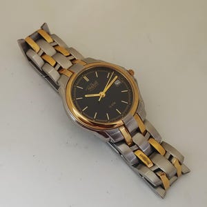 K&ouml;nnte beinhalten: Eine Armbanduhr mit schwarzem Zifferblatt, goldfarbenen Zeigern und L&uuml;nette sowie einem silber- und goldfarbenen Metallarmband. Das Zifferblatt zeigt das Datum und den Markennamen. Ein klassisches Accessoire.