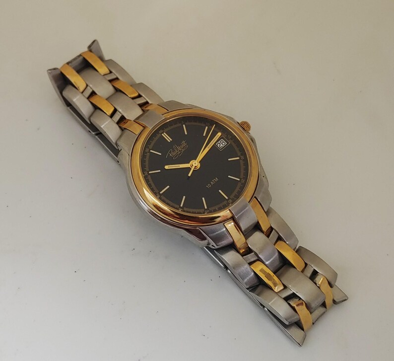 K&ouml;nnte beinhalten: Eine Armbanduhr mit schwarzem Zifferblatt, goldfarbenen Zeigern und L&uuml;nette sowie einem silber- und goldfarbenen Metallarmband. Das Zifferblatt zeigt das Datum und den Markennamen. Ein klassisches Accessoire.