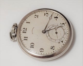 Puede incluir: Un reloj de bolsillo plateado con una esfera blanca y marcadores de hora romanos negros. El reloj tiene un pequeño dial de segundero en la posición de las 6 en punto. La esfera del reloj dice "Illinois".