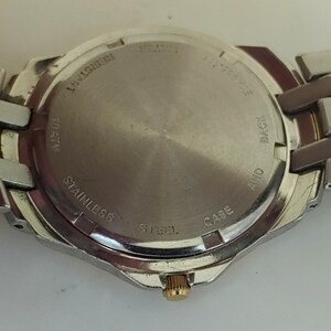 K&ouml;nnte beinhalten: Edelstahl-Uhrenr&uuml;ckseite mit silbernem Armband. Auf der Uhrenr&uuml;ckseite stehen die W&ouml;rter &bdquo;RESISTANT&ldquo;, &bdquo;STAINLESS STEEL&ldquo;, &bdquo;10 ATM&ldquo; und &bdquo;CASE AND BACK&ldquo;. Die Uhr hat eine goldfarbene Krone.