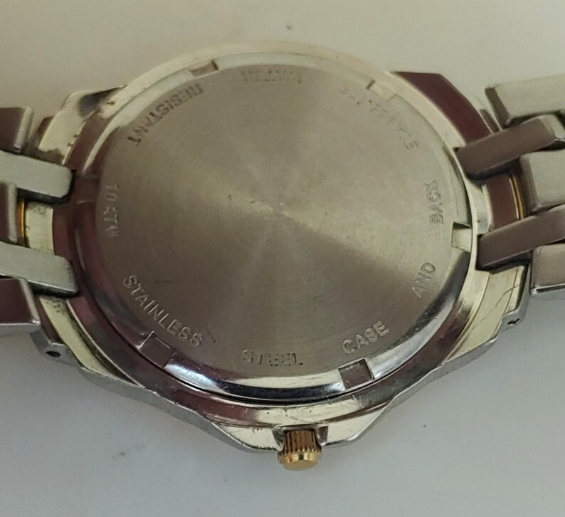 K&ouml;nnte beinhalten: Edelstahl-Uhrenr&uuml;ckseite mit silbernem Armband. Auf der Uhrenr&uuml;ckseite stehen die W&ouml;rter &bdquo;RESISTANT&ldquo;, &bdquo;STAINLESS STEEL&ldquo;, &bdquo;10 ATM&ldquo; und &bdquo;CASE AND BACK&ldquo;. Die Uhr hat eine goldfarbene Krone.
