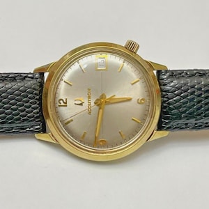 May include: A gold-toned Accutron wristwatch with a silver face, gold hands, and a black leather band. The watch features a date window at the 3 o'clock position and the word "ACCUTRON" printed on the face. The watch band is approximately 0.75 inches wide.