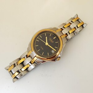 K&ouml;nnte beinhalten: Eine gold- und silberfarbene Armbanduhr mit schwarzem Zifferblatt. Die Uhr hat goldene Zeiger und Stundenmarkierungen, eine Datumsanzeige und ein zweifarbiges Metallarmband. Auf dem Zifferblatt steht "10 ATM".