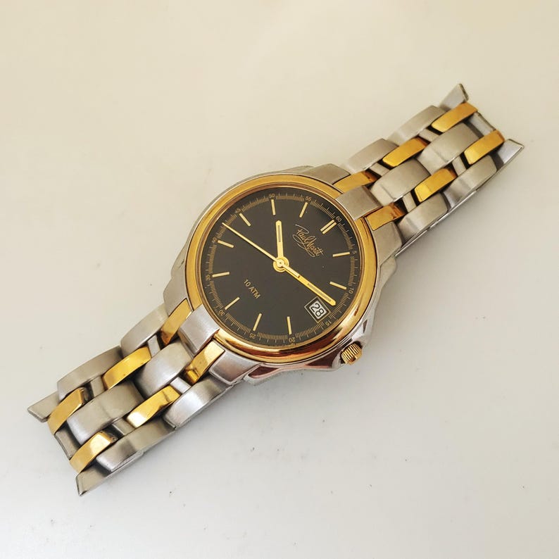 K&ouml;nnte beinhalten: Eine gold- und silberfarbene Armbanduhr mit schwarzem Zifferblatt. Die Uhr hat goldene Zeiger und Stundenmarkierungen, eine Datumsanzeige und ein zweifarbiges Metallarmband. Auf dem Zifferblatt steht "10 ATM".