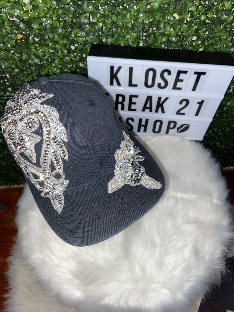 May include: A black baseball cap with silver rhinestone embellishments. The cap has a curved brim and a mesh back. The rhinestones are arranged in a floral design on the front of the cap.