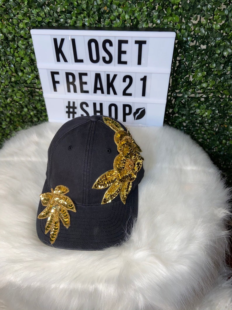 Puede incluir: Gorra de b&eacute;isbol negra con adornos de lentejuelas doradas en forma de hojas. La gorra tiene una visera curvada y un fondo blanco.