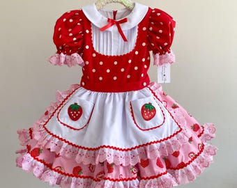 Op de afbeelding: Een rood-witte polkadotjurk met een witte kraag en een rode strik. De jurk heeft een witte schort met aardbeienzaken en een roze rok met aardbeien- en kanten details. De jurk hangt aan een hanger.