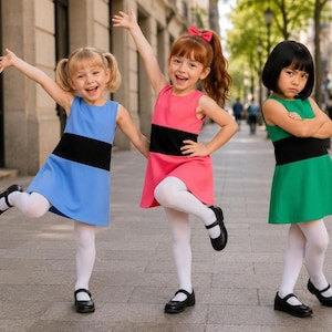 Pode incluir: Três meninas posam com vestidos coloridos numa rua da cidade. Cada uma usa um vestido sem mangas com uma faixa preta na cintura. Os vestidos são azuis, rosa e verdes. Elas usam meias brancas e sapatos pretos.