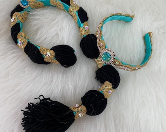 May include: An ornate, decorative hair accessory set. The set features black yarn accents, gold trim, and teal fabric. Sparkling gemstones and a tassel complete the design. The set is designed to be worn as a hair accessory.