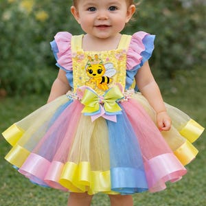 May include: A young child in a vibrant tutu dress with a bee motif. The dress has a yellow top with a bee illustration, a rainbow skirt, and a matching headband with a bow. The child is also wearing white shoes.
