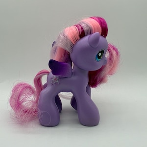 Könnte beinhalten: Ein lila Spielzeugpony mit einer rosa und weißen Mähne und einem Schweif. Das Pony hat blaue Augen, ein sternförmiges Schönheitszeichen und lila Flügel. Das Spielzeug steht auf einer weißen Oberfläche.