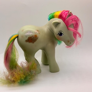 Può includere: Un pony giocattolo verde chiaro con criniera e coda arcobaleno. Il pony ha una criniera rosa e arcobaleno, una coda arcobaleno e macchie glitterate sul lato. Il pony è in piedi su tutte e quattro le zampe.