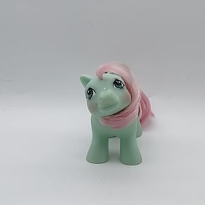 May include: A vintage toy pony in a standing position. The pony has a light green body, pink hair, and blue eyes. The toy is a collectible item, likely from the 1980s or 1990s. The pony is in good condition and appears to be well-preserved.