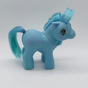 May include: A light blue toy pony with a blue mane and tail. The pony has green eyes and pink star-shaped markings on its side. The pony is standing on all four legs.