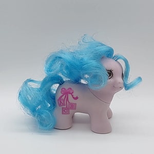 May include: A small, light purple pony with a blue curly mane and tail. The pony has a pink bow and a design of three blocks with the letters A, B, and C on its side.