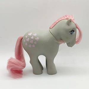May include: A gray toy pony with pink hair and tail. The pony has a cluster of pink hearts on its flank. The pony's eyes are blue. The mane and tail are a vibrant pink color. This vintage toy is a collectible item.
