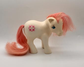 May include: A vintage toy pony in off-white with a pink mane and tail. The pony has blue eyes and a pink heart-shaped design on its side. The toy is standing on all four legs against a white background.