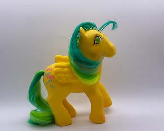 May include: A yellow toy pony with green and blue hair and tail. The pony has wings and a design on its flank. The pony has green eyes and is standing on all four legs. The pony is a vintage toy.