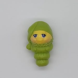Könnte beinhalten: Eine kleine, geformte Kunststofffigur eines Raupencharakters mit grüner Schlafmütze. Die Figur hat ein gelbes Gesicht, große blaue Augen und einen segmentierten grünen Körper mit einem gerüschten Kragendetail um den Halsbereich im Cartoon-Stil.