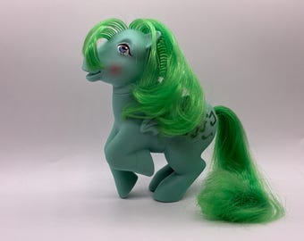 May include: A vintage teal-colored toy pony with bright green hair and tail. The pony has a pink blush on its cheeks and a decorative design on its flank. The pony is a collectible toy.