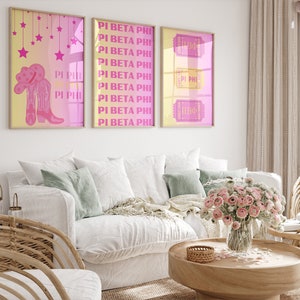 May include: Three framed prints with pink and yellow backgrounds. The prints feature the text "Pi Beta Phi" and "Pi Phi" in pink and yellow letters. The left print features a pair of pink cowboy boots with a pink cowboy hat. The middle print features the text "Pi Beta Phi" repeated multiple times. The right print features the text "Pi Phi" and "ПВФ" in pink letters.